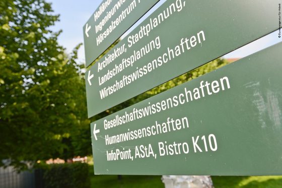 Drei Wegweiser an einem Unicampus weisen zu verschiedenen Hörsälen und Fakultäten.